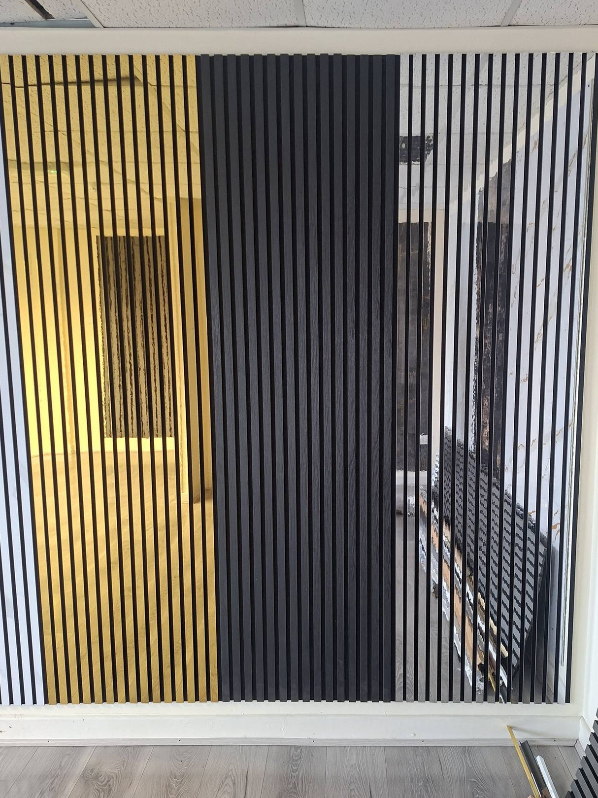 A modern wall-mounted slatted screen or partition panel with three vertical sections: white, black, and gold, installed in an indoor space with wood-look flooring.