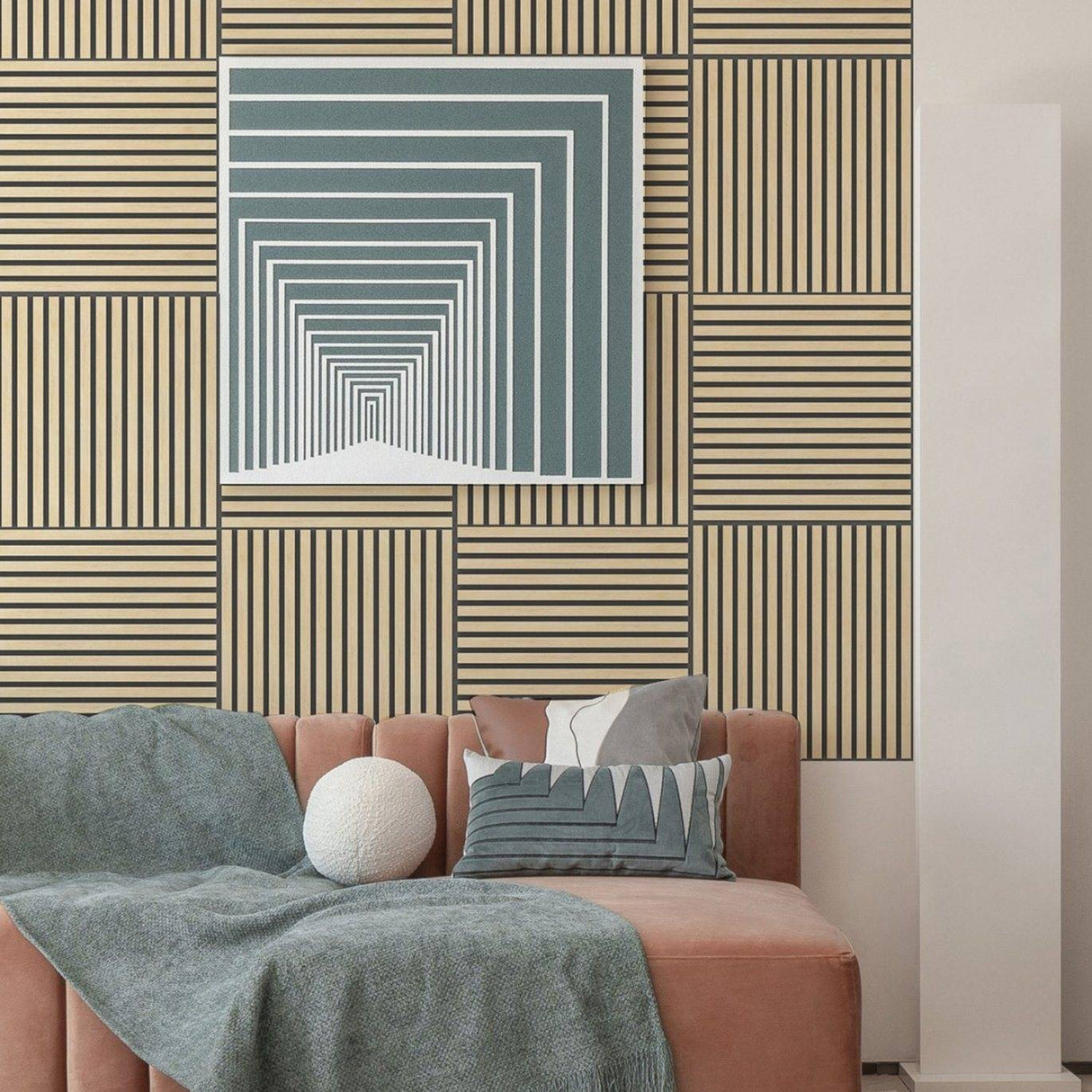 A modern living room scene featuring a geometric wall art print with concentric arches in slate blue and white, mounted on a wall with beige and brown striped panels. Below the artwork is a blush pink sofa styled with a textured blue throw blanket, a white spherical pillow, and patterned accent pillows.