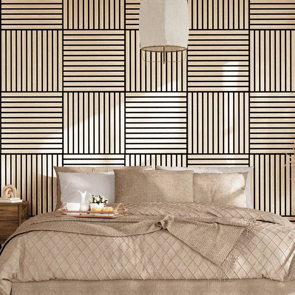 A styled bedroom scene featuring a bed dressed in beige quilted bedding and pillows, set against a bold geometric wallpaper with black and beige stripes. A matching patterned rug covers the floor, and a wooden nightstand with a lamp is visible on the left.