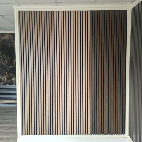 A vertical slatted wall panel in a warm medium brown finish, mounted within a white frame on a ceiling and floor, with a tiled ceiling visible above.