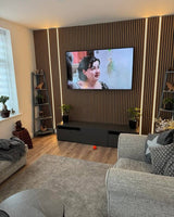 A modern living room setup featuring a large wall-mounted TV displaying a woman, set against a brown wood slat accent wall with vertical LED strip lighting. The room includes a gray fabric sofa, a matching armchair, a black media console, and a light-colored area rug.