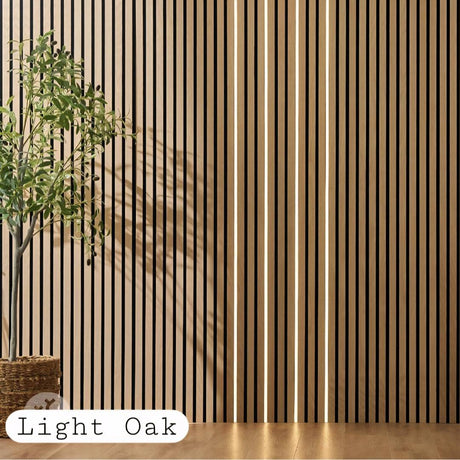 Light oak vertical wood slat wall panel with integrated warm LED strip lighting, shown in a styled interior setting with a potted plant and a floor label reading 'Light Oak'.