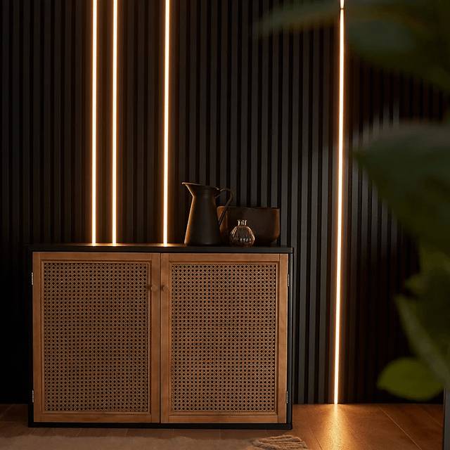 A modern wooden sideboard cabinet with two woven cane doors, styled with dark ceramic vessels against a dark wood slatted wall with vertical LED light strips.