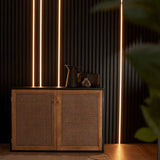 A modern wooden sideboard cabinet with two woven cane doors, styled with dark ceramic vessels against a dark wood slatted wall with vertical LED light strips.
