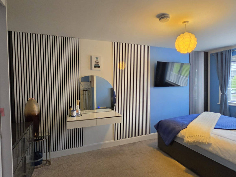A modern bedroom interior with a bed dressed in blue and white bedding, a wall-mounted TV, a yellow globe pendant light, striped wall panels, and a blue accent wall.